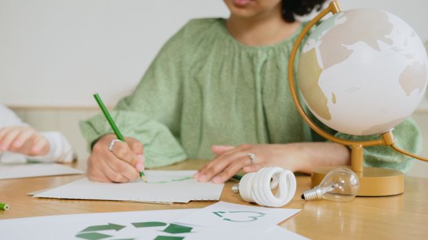 Person drawing recycling symbol at desk with globe and eco-friendly items.